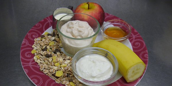 Müslischaum, arrangiert mit den darin enthaltenen Zutaten: Banane, Getreideflocken, Apfel und Joghurt.