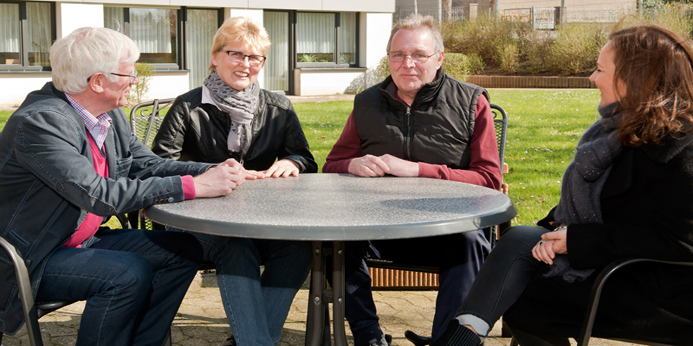 Bewohner sitzen mit ihren Angehörigen im Garten am Tisch.