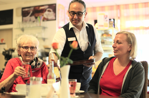 Mitarbeiter serviert einer Bewohnerin und ihren Angehörigen Getränke in der Cafeteria des Seniorencentrums.